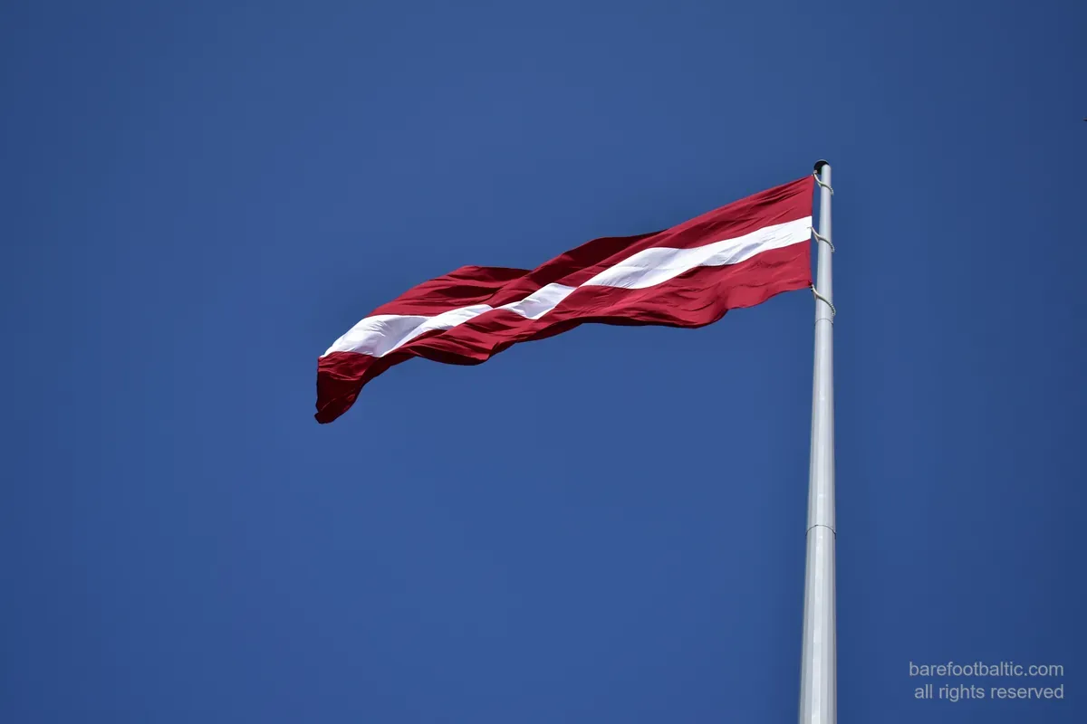 Lettlands flagga, karminröd med ett vitt band, vajande mot en blå himmel.
