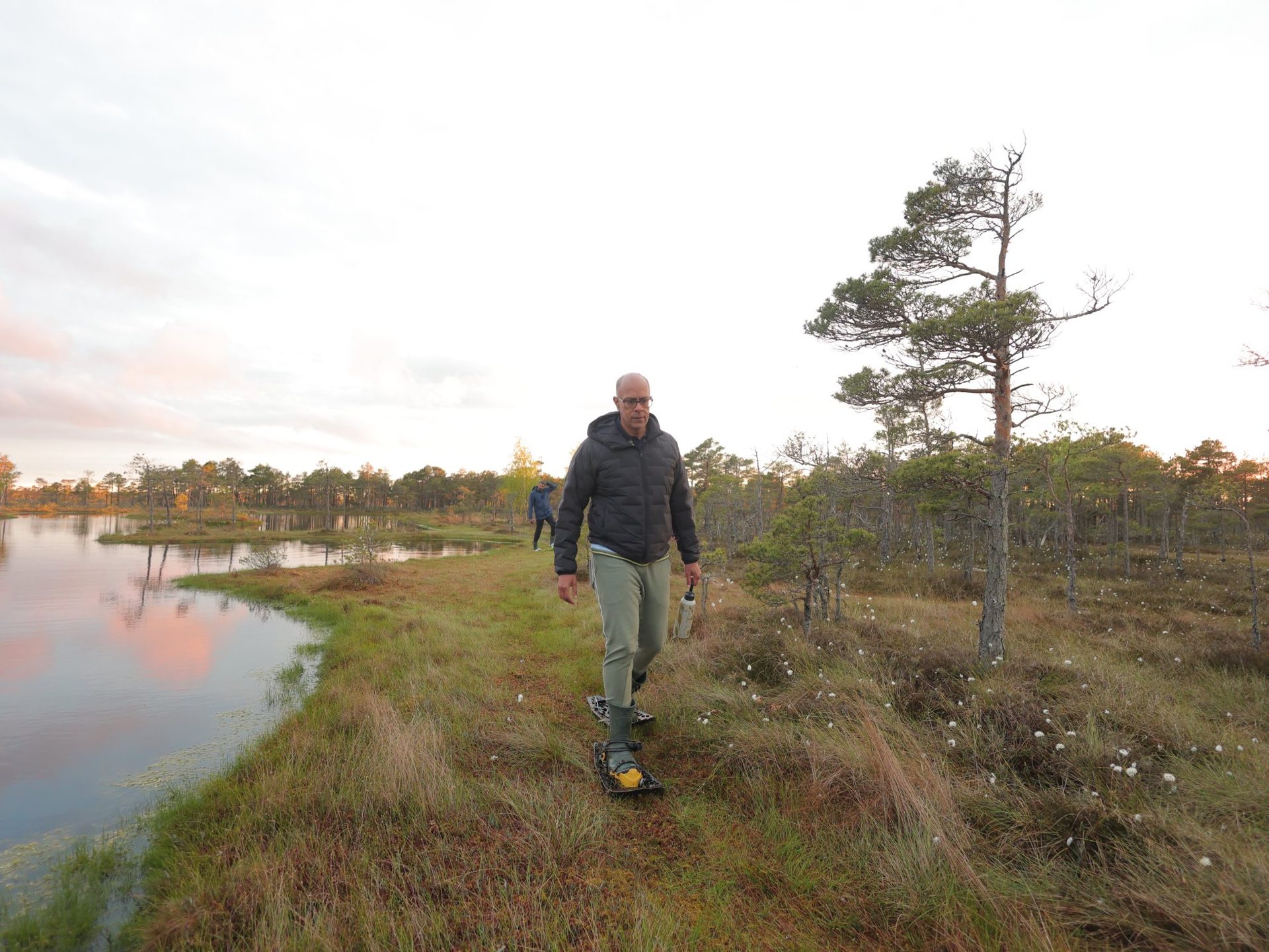 Ensam vandrare med myrskor intill en göl som speglar gryningens rosa ljus
