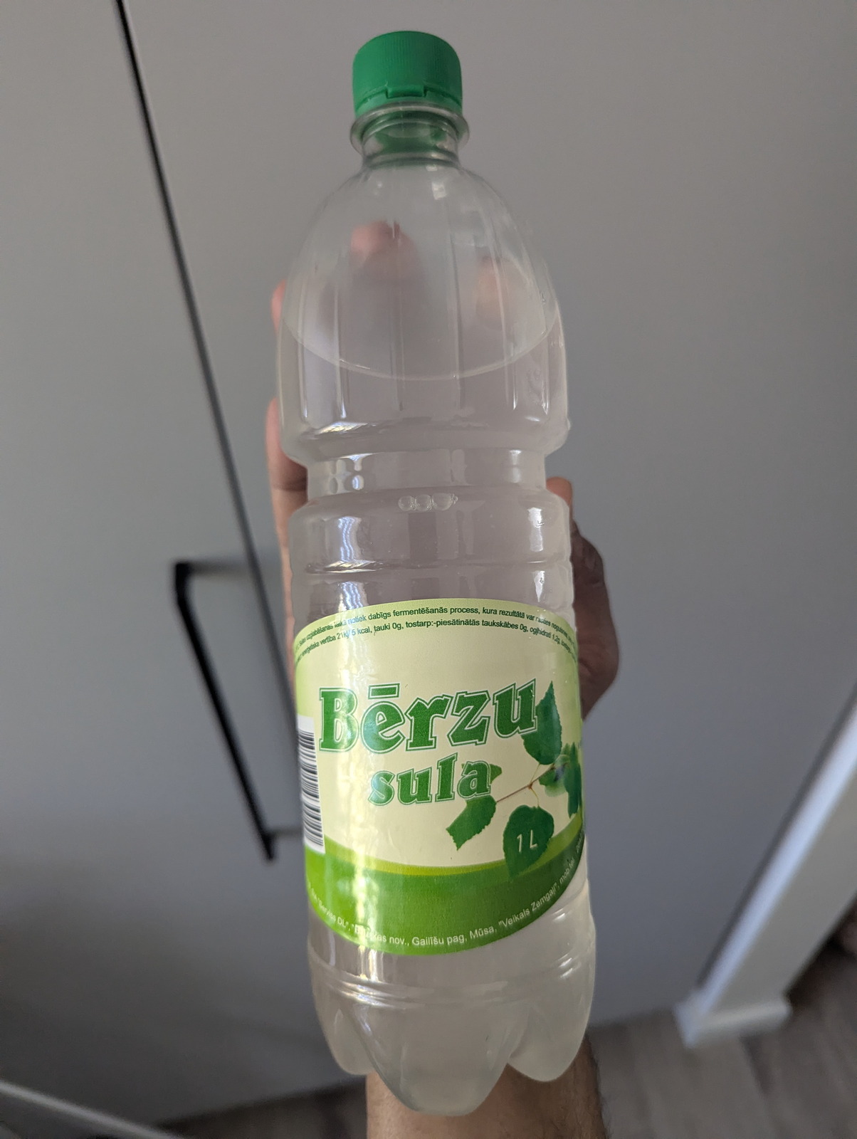 En literflaska av PET med Bērzu sula (lettisk björksav) med en grön lövmönstrad etikett, hållen upp mot en köksvägg