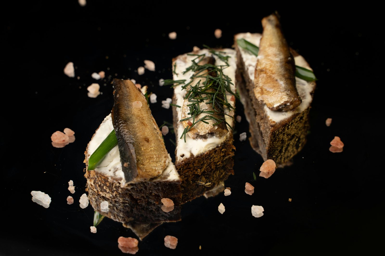 Tre rökta skarpsillar på rågbröd med gräddfil och dill, mot svart bakgrund